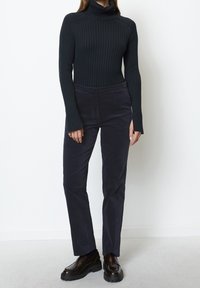 Musta ribbineulottu poolopaita, jossa pitkät hihat, yhdistetty tummansinisiin samettihousuihin. Kengät ovat tummanruskeat liukusäädettävät, paksulla pohjalla.