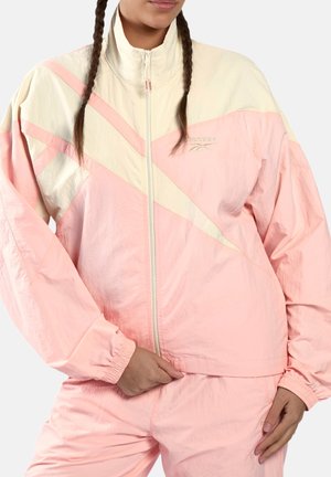 Frau mit zwei Zöpfen, die einen rosa-weißen Reebok-Trainingsanzug trägt und mit einer Hand den Saum der Jacke hält.