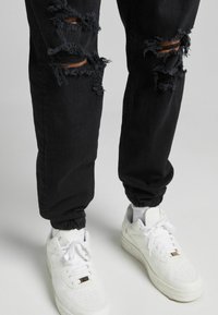 Ben klädda i svarta rivna jeans med elastiska avslut och vita sneakers mot en enkel ljus bakgrund.