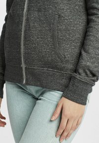 Charcoal šedá mikina na zips s textúrovanou látkou, prednými vreckami a rebrovanými manžetami. V kombinácii so svetlo modrými, priliehavými denimovými džínami.