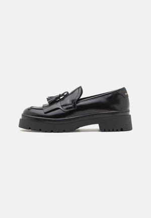 Black patent leather loafers with a chunky rubber sole, featuring a distinctive tassel detail and scalloped edges.