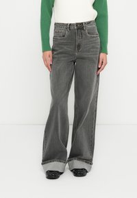 Sivá džínsy s vysokým pásom a širokými nohavicami s bledým efektom. Majú predné vrecká, viditeľné švy a ohrnuté lemovanie. Skombinované so zeleno-bielym topom.