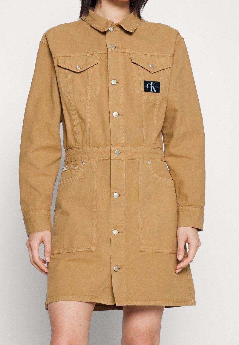Femme portant une robe boutonnée beige longueur genou avec des poches poitrine et un patch logo Calvin Klein noir sur le côté gauche.
