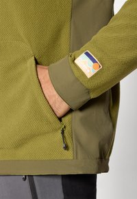 Giacca verde con zip, realizzata in tessuto testurizzato e maniche lisce. Presenta un'etichetta con logo sul polsino e una tasca in vita.