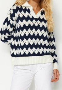 Donna che indossa un maglione lavorato a maglia a motivi di diamante blu navy e bianco con colletto bianco e pantaloni bianchi, in piedi con una mano sui fianchi.