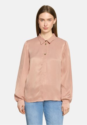 Frau trägt eine hellrosa satinierte Bluse mit langen Ärmeln und goldenen Knöpfen, kombiniert mit hellblauen Jeans, steht vor einem schlichten Hintergrund.