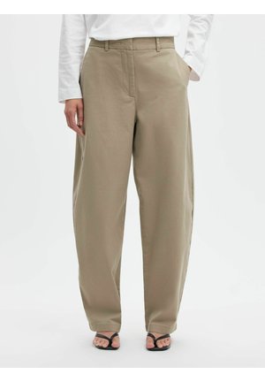 Løse beige bukser med sidelommer, båret med en hvid langærmet top og sorte åbne sandaler på en ensfarvet baggrund.
