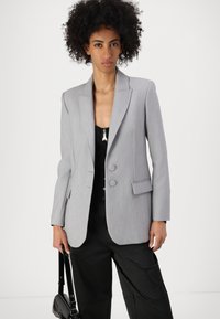 Lys grå skreddersydd blazer med skrårever, to knapper og sidelomme. Brukt over en svart topp, kombinert med svarte bukser og håndveske.
