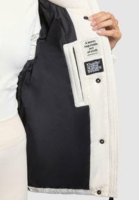 Witte pufferjas met een zwarte voering. Voorzien van twee stoffen labels en drukknoopsluitingen. De binnenvoering is soepel met een matte afwerking.
