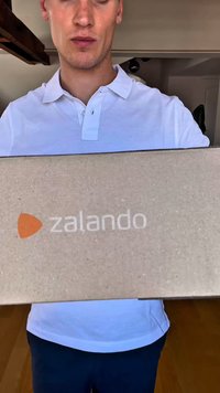 Rjava kartonska škatla z logo "zalando" v beli in oranžni barvi, ki jo drži oseba v svetlem polo majici.