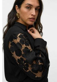 Blusa nera con maniche in pizzo floreale a motivi texture. Design con colletto e dettaglio dei bottoni, corpo in tessuto liscio.