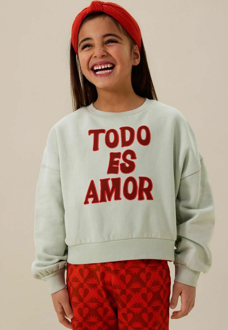 Sweat à capuche vert clair avec le texte brodé rouge "TODO ES AMOR", associé à un pantalon rouge à motifs coeurs. Design confortable et décontracté.