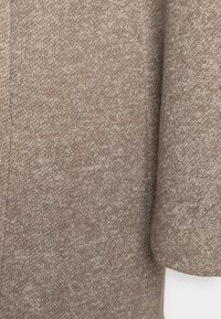 Vaaleanruskea neulottu kangas, jossa on kirjavuutta. Kuvassa korostuu materiaalin kudontakuviot ja pehmeys.