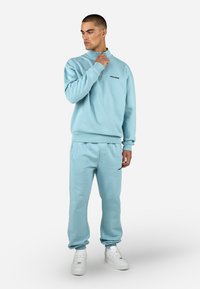 Gaiši zila džemperis ar puszipsa apkakli un melnu logotipu, savienots ar atbilstošiem sporta biksēm ar sānu kabatām un elastīgiem aproces. Baltas kedas.