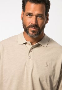 Béžové polo tričko vyrobené z měkké tkaniny, s upraveným límečkem, plaketou na tři knoflíky a vyšitým logem na hrudi.