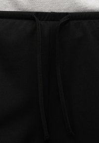 Μαύρο παντελόνι από ύφασμα με κορδόνι στη μέση, εμφανίζοντας το επάνω μπροστινό μέρος με δύο χαλαρά άκρα κορδονιών.