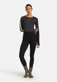 Kvinna som står i en svart och grå långärmad topp och leggings med beige ränder, bär stickade grå strumpor mot en vit bakgrund.
