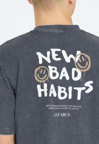 Henkilö, jolla on yllä haalistunut musta T-paita, jossa lukee "NEW BAD HABITS", kaksi vaaleaa hymyilevää kasvua ja pieni määritelmäteksti takana.