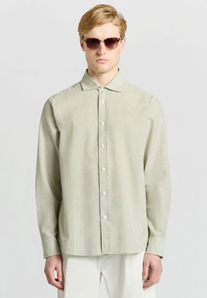 Uomo biondo che indossa occhiali da sole tartarugati, camicia a righe verde chiaro con bottoni e pantaloni bianchi, in piedi davanti a uno sfondo chiaro e semplice.