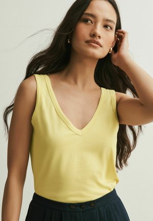 Frau mit langem dunklem Haar, die goldene Creolen, ein gelbes ärmelloses V-Ausschnitt-Oberteil und dunkle hochtaillierte Hosen trägt, während sie mit einer Hand ihr Haar richtet.