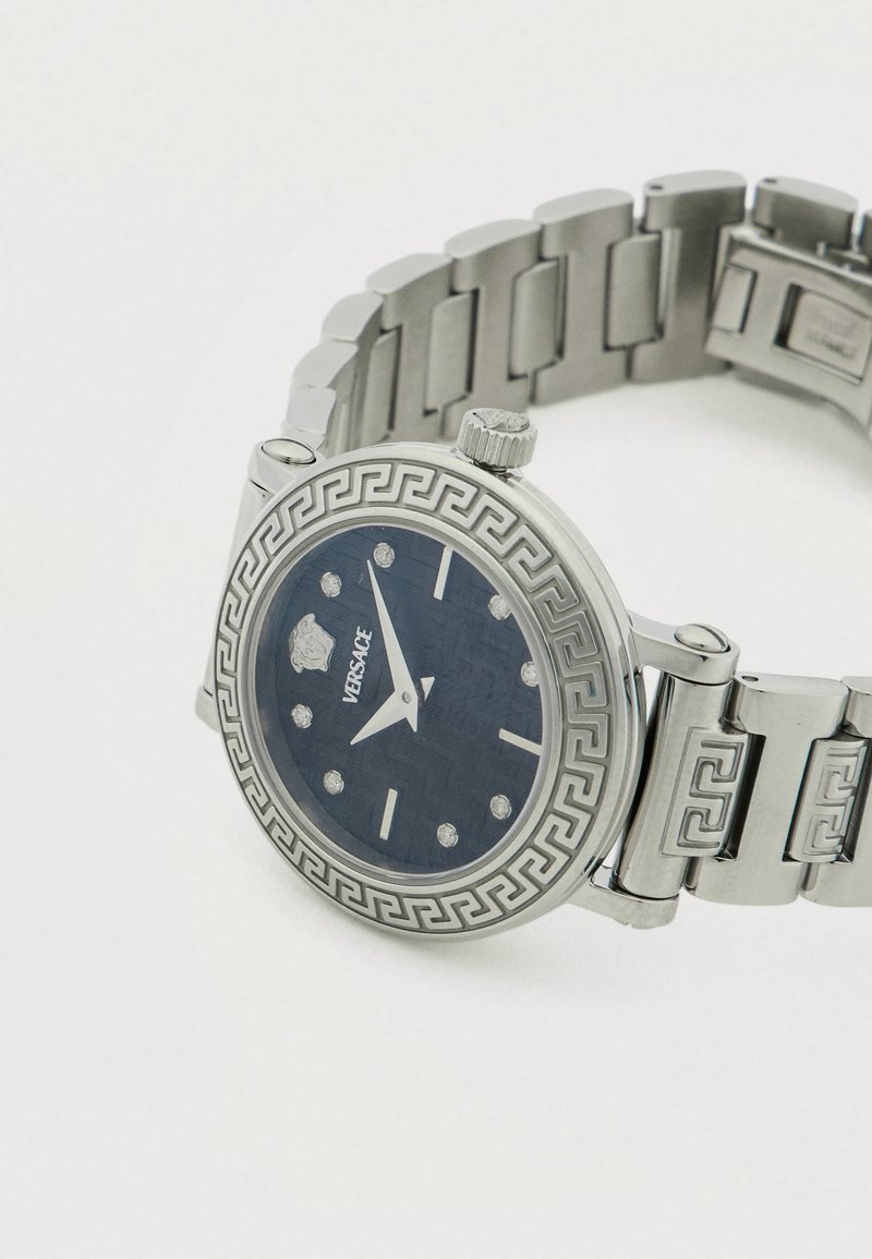 Silberne Uhr aus Edelstahl mit einem marineblauen, strukturierten Zifferblatt, Diamant-Stundenmarkierungen und einer Lünette mit griechischem Schlüssel-Muster. Metallarmband mit Gliedern.