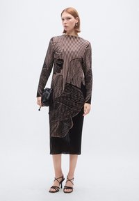 Robe rayée noire et nude avec texture côtelée, longueur mi-mollet, manches longues, motif abstrait, associée à un sac à main noir et à des talons à brides.