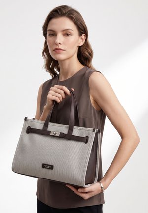 Femme tenant un sac à main texturé beige et marron avec deux poignées, portant un top marron sans manches et un bracelet en argent, sur un fond uni.