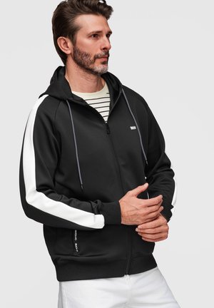 Homme portant un sweat à capuche noir zippé avec des rayures blanches sur les manches et une chemise blanche rayée, debout devant un fond clair uni.