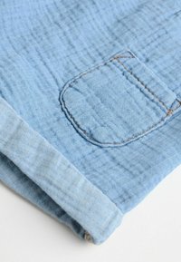 Helles Blau, strukturiertes Gewebe mit sichtbaren Nähten; verfügt über eine aufgesetzte Tasche und einen umgeschlagenen Saum. Das Material scheint weich und atmungsaktiv zu sein.