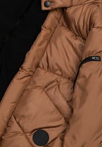 Braune Pufferjacke mit glatter Textur, schwarzen Akzenten, einer Reißverschlusstasche und einem runden Logo-Patch auf der linken Seite.