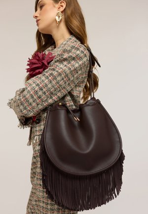 Mujer con chaqueta de cuadros con flor de tela burdeos sostiene un gran bolso de cuero marrón oscuro con flecos en el hombro.