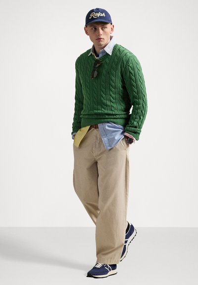 Green cable-knit sweater, light blue shirt, yellow tie, beige pants, and blue sneakers. Navy cap with "Ralph" logo. Sunglasses on shirt.
