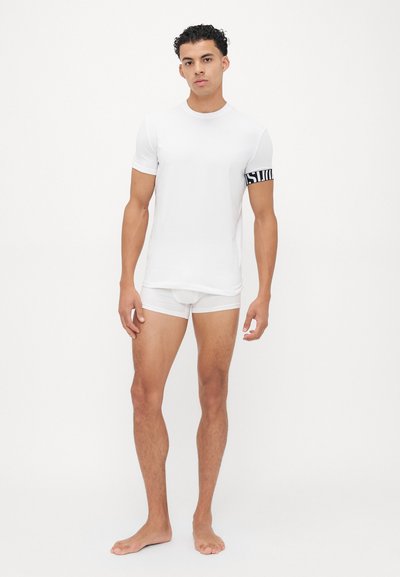 Weißes kurzärmliges T-Shirt mit schwarfer Grafik auf dem linken Arm; getragen mit weißen Boxer-Shorts. Glattes, leichtes Material; taillierte Passform.