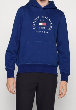 Mann trägt einen blauen Tommy Hilfiger Hoodie mit Fronttasche und Logo mit Flagge sowie dem Text "Tommy Hilfiger EST 1985 New York".