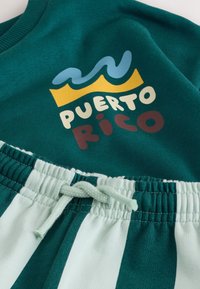 Grüner Sweatshirt mit gelbem und blauem Wellenmuster und dem Schriftzug „Puerto Rico“, kombiniert mit grün-weiß gestreiften Jogginghosen mit Kordelzug.