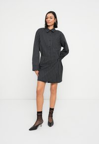 Φόρεμα πουκάμισο με μαύρες ρίγες, μακριά μανίκια, γιακά, κουμπιά στο μπροστινό μέρος και λεπτομέρεια με κόμπο στο πλάι. Φορεμένο με διάφανα μαύρα καλτσάκια και τακούνια.