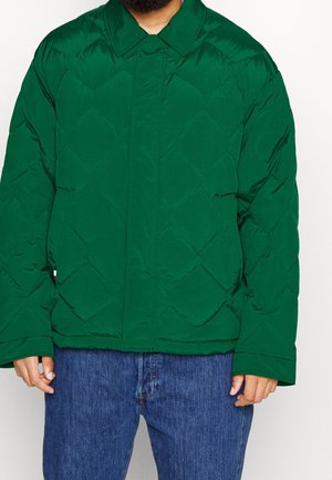 Green quilted puffer jacket with a classic collar, diamond pattern, and a relaxed fit; worn over blue denim jeans.
