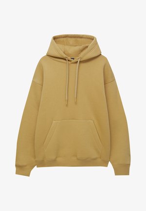 Person trägt senfgelbe Kapuzenjacke, übergroße hellblaue Jeans und weiße Turnschuhe und steht mit dem Rücken zur Kamera vor weißem Hintergrund.