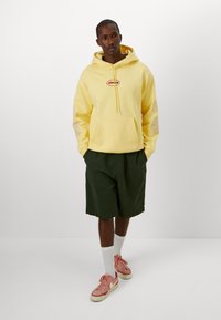 Hooded yellow sweatshirt with a logo, large front pocket, light fabric, paired with dark green shorts and pink sneakers. White socks visible.