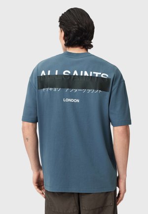 Tummatukkainen mies, jolla on yllään sininen ylipitkä t-paita, jossa on valkoinen teksti ja musta teippi takana, seisoo yksinkertaisen taustan edessä.