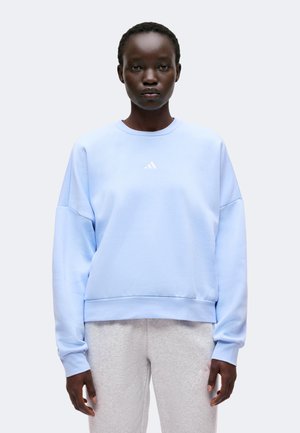 Jeune personne aux cheveux courts portant un sweat-shirt bleu clair à manches longues et un pantalon de jogging gris clair, debout devant un fond uni.