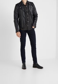 Chaqueta de motorista de cuero negro, camisa negra con patrón, jeans oscuros y botas negras hasta el tobillo. La chaqueta cuenta con cremalleras y hombreras.