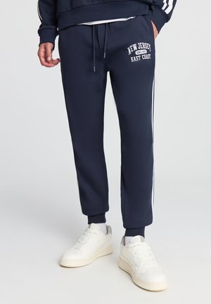 Pantaloni tuta blu con strisce laterali bianche e testo "New Jersey East Coast". Presenta una vita elasticizzata con coulisse e polsini affusolati.