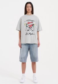 Grijze oversized t-shirt met een schedelafbeelding en een kleurrijk hartontwerp, gecombineerd met lichtblauwe denimshorts en witte sneakers.