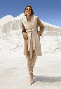 Vrouw die glimlacht en buiten loopt, gekleed in een beige jasje met riem, een coltrui, een op maat gemaakte broek en enkelboots tegen een witte rotsachtige achtergrond.