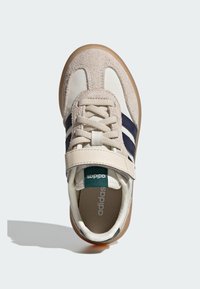 Zapatillas con parte superior de ante beige y sintético. Presentan rayas azules y blancas, una correa de Velcro y una suela de goma. Amortiguación interna visible.