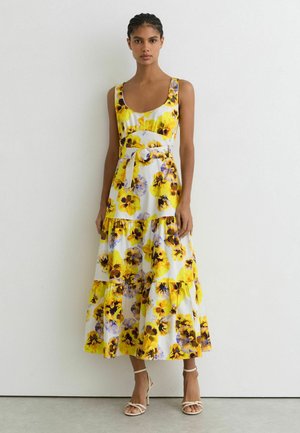 Kobieta w białej sukience midi bez rękawów z żółto-fioletowym kwiatowym wzorem, z paskiem w talii i białych sandałkach na paski, stojąca przy białej ścianie.