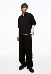 Ung mand iført en sort kortærmet skjorte med knapper over en hvid langærmet top, brede sorte bukser og sorte snørebåndssko, stående.