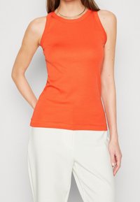 Top senza maniche in cotone arancione con scollatura rotonda, design aderente e texture liscia. Abbinato a pantaloni bianchi.