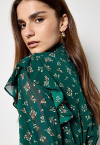 Luźna zielona bluzka w kwiatowy wzór, z falbanami na rękawach i marszczonym dekoltem, wykonana z lekkiego materiału o gładkiej fakturze.